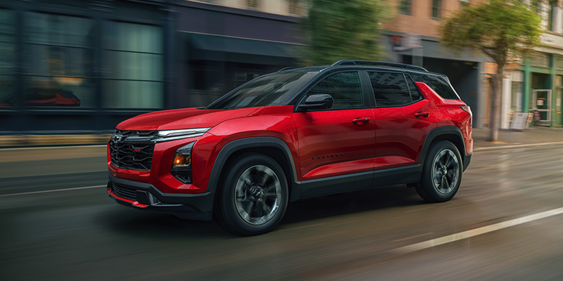 Red 2026 Chevrolet Equinox SUV driving through a city street, shown in a front three-quarter view with motion blur in the background, black roof and trim, and modern LED headlights.